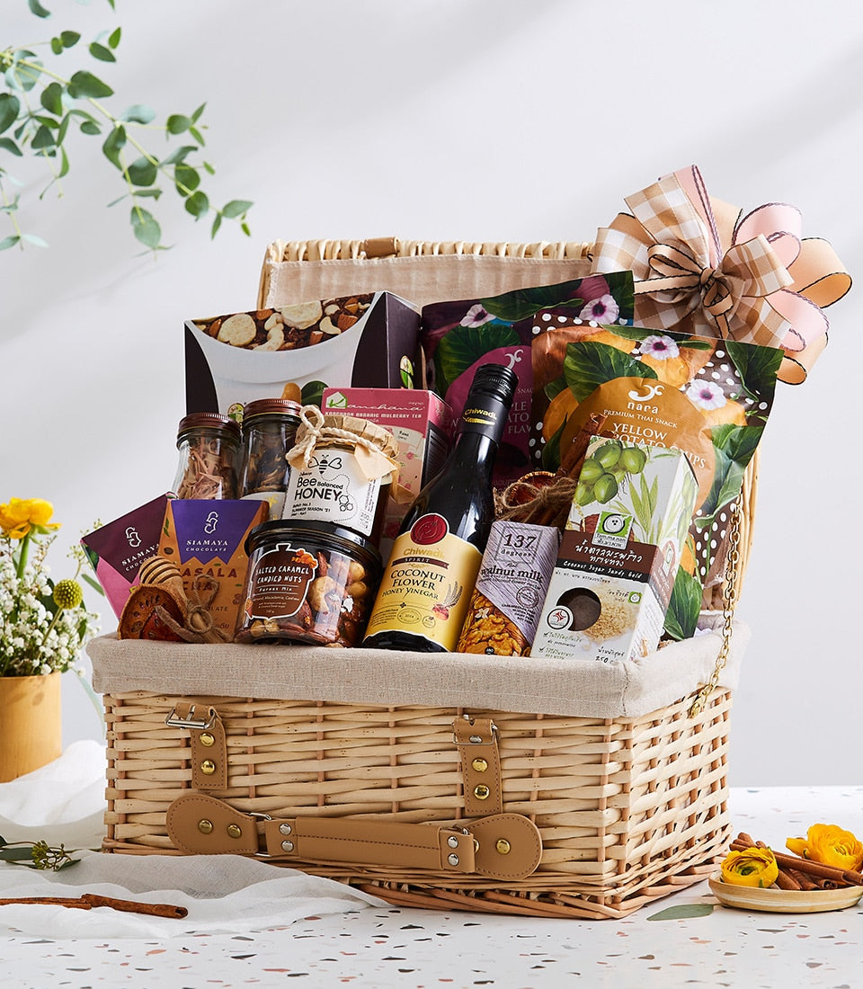 Light brown wicker picnic basket filled with Thai organic gourmet foods, nuts, honey, and snacks, tied with checkered ribbon