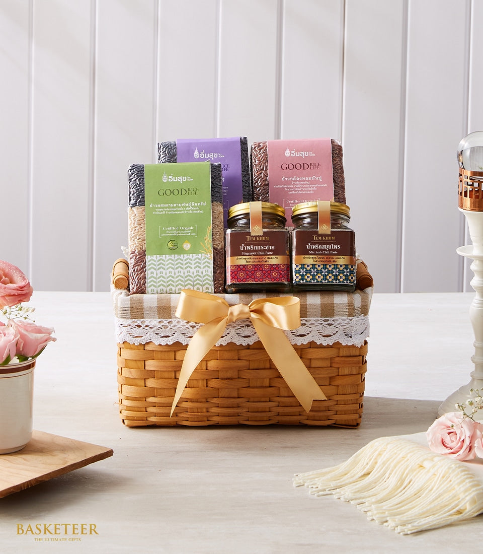 Woven gift basket with Thai rice varieties, artisan chili paste jars, pink roses, and candle in gold-accented presentation