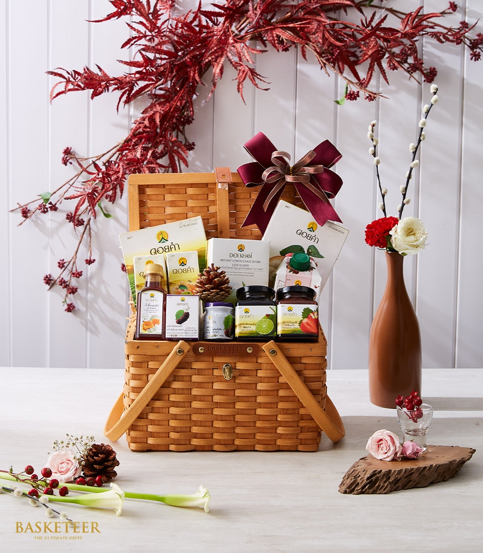 Woven wicker picnic basket gift hamper with gourmet preserves, lemongrass drink, and burgundy ribbon, styled with white