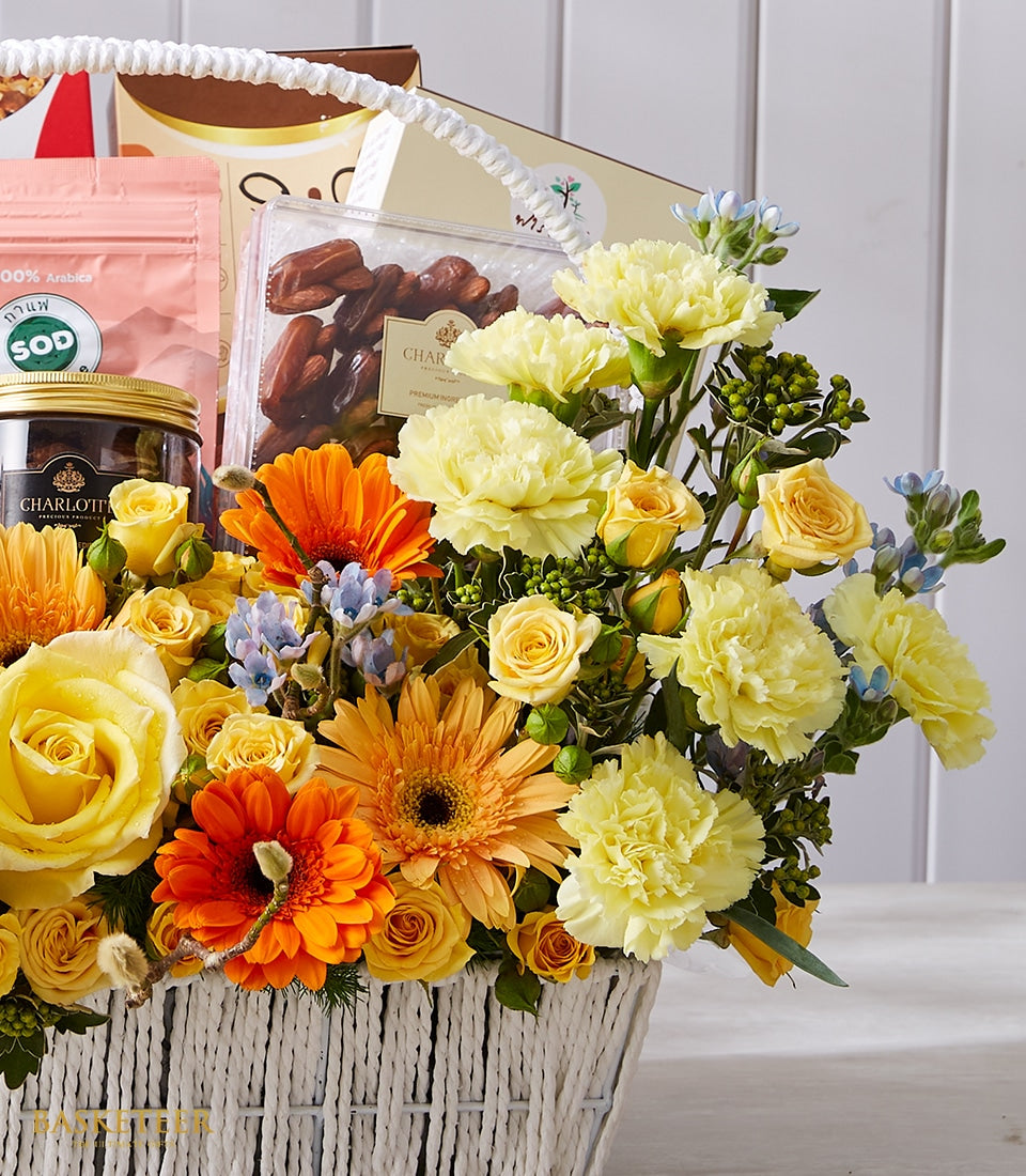 Luxury gift basket with yellow, orange and blue fresh flowers, gourmet treats and premium beverages in white woven basket