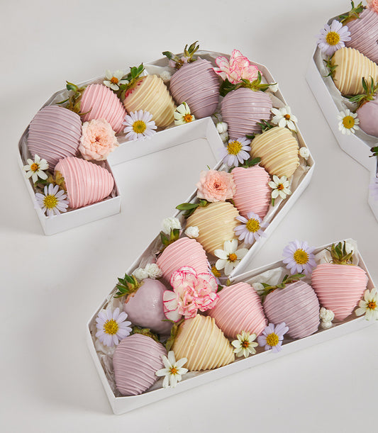 Chocolate-dipped strawberry number box with pastel pink and purple coatings, fresh flowers, and greenery for birthday
