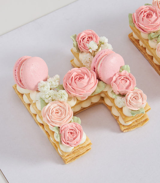 Monogram letter H pastry cake with cream layers, pink buttercream roses, and macarons for birthday celebrations