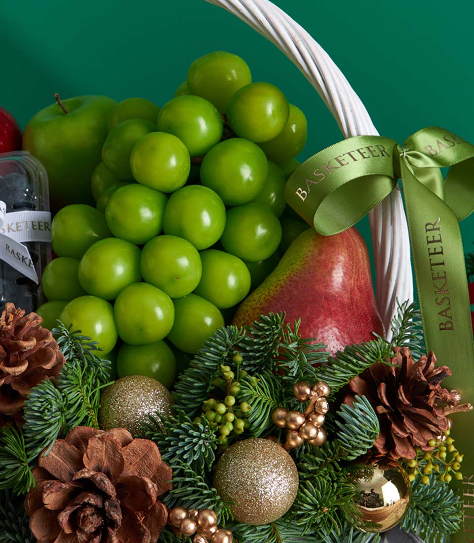 White woven Christmas fruit basket with green grapes, apple, pear, and gold ornaments, festive holiday gift hamper