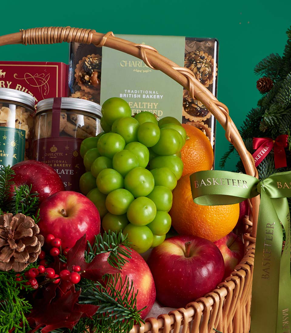 Premium woven basket filled with fresh green grapes, red apples, oranges, artisan cookies, and festive pine garland with red