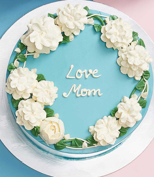 Light blue frosted birthday cake with "Love Mom" message, white buttercream flowers and green leaves