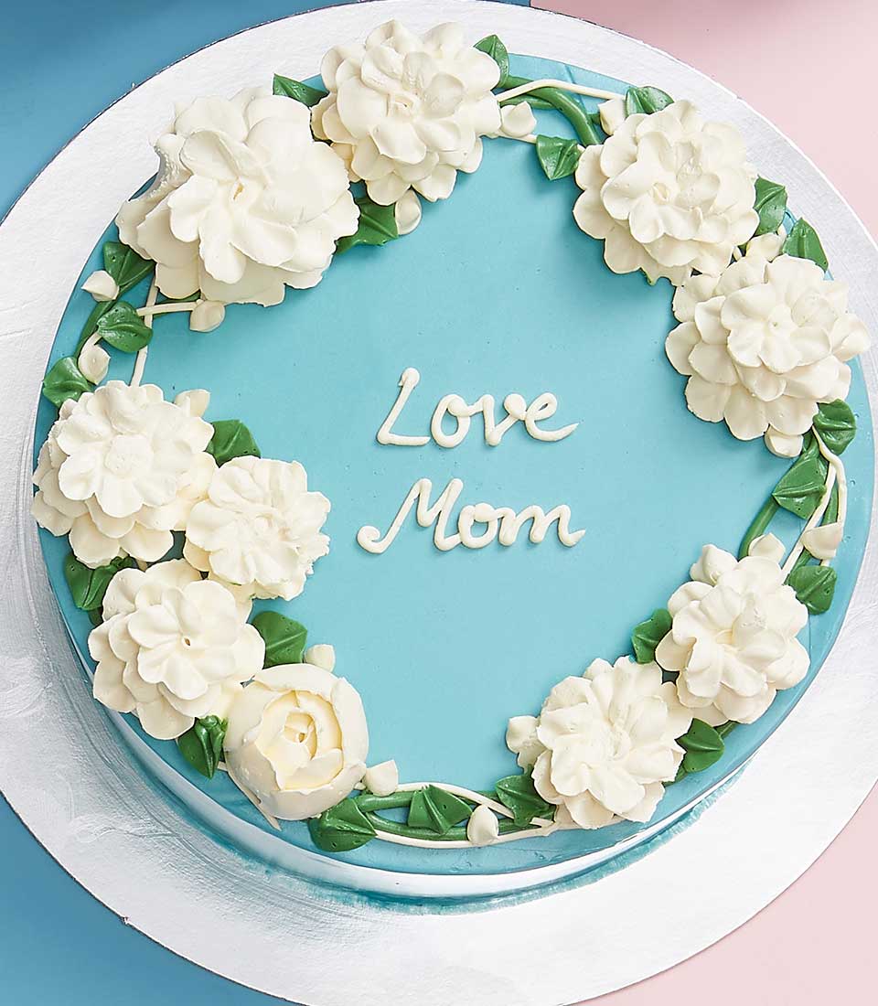 Light blue frosted birthday cake with "Love Mom" message, white buttercream flowers and green leaves
