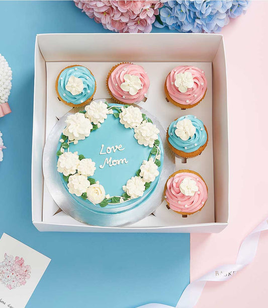 Light blue cake with white piped flowers and "Love Mom" message, surrounded by blue and pink frosted cupcakes with pearl
