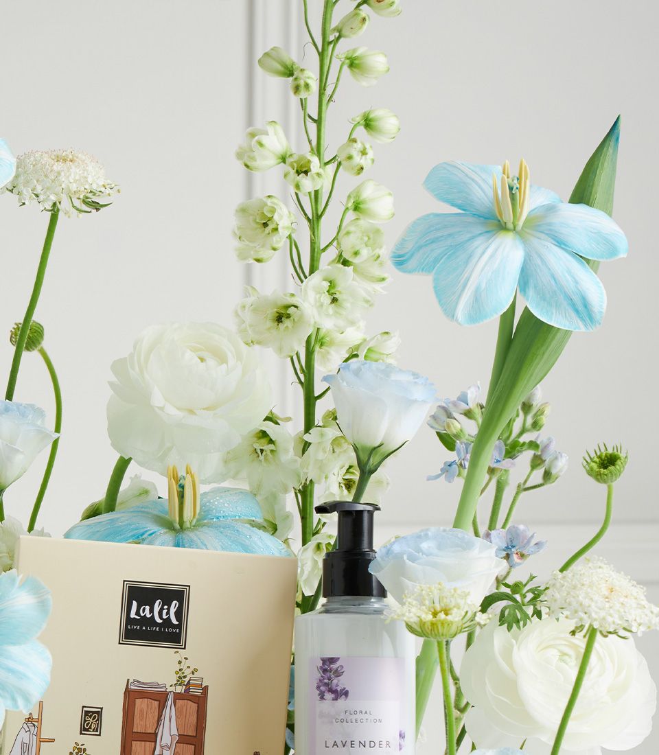 Luxury spa gift basket with lavender products, white ranunculus, blue delphiniums and roses for Mother's Day pampering