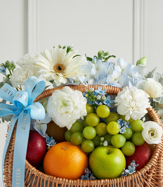 Wicker fruit and flower gift basket with fresh apples, oranges, grapes, white gerberas, ranunculus, and blue hydrangeas for