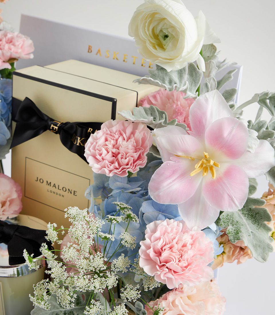 Luxury Jo Malone candle surrounded by pastel pink carnations, white ranunculus, and blue hydrangeas in Mother's Day gift