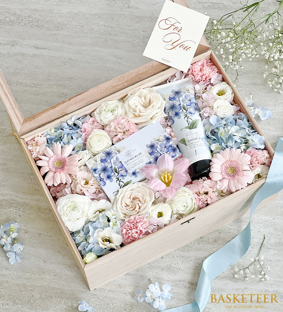 Luxury wooden gift box with fresh pink roses, hydrangeas, and ranunculus paired with blue floral spa products for Mother's
