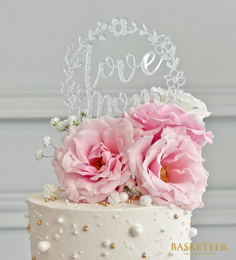 Two-tier vanilla cake with pink roses, white frosting, and silver "Love Mom" topper for Mother's Day