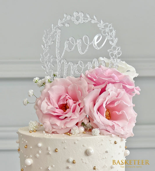 Two-tier vanilla cake with pink roses, white frosting, and silver "Love Mom" topper for Mother's Day