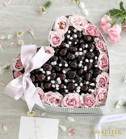 Heart-shaped gift box with dark cherries, pink roses, and baby's breath flowers tied with pink satin ribbon