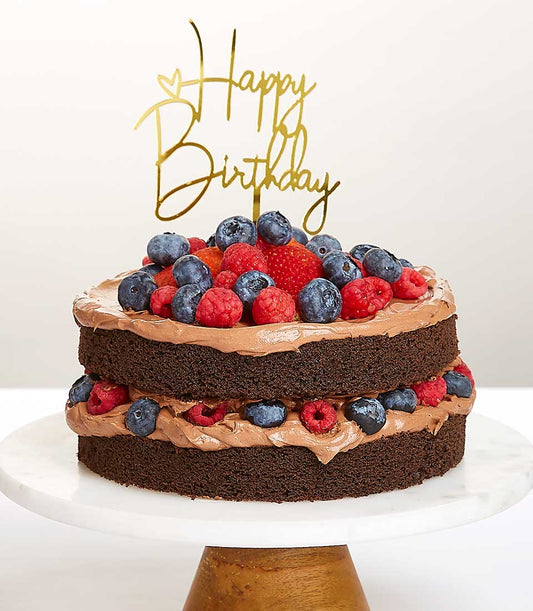 Two-tier chocolate cake with berries and gold Happy Birthday topper on marble stand