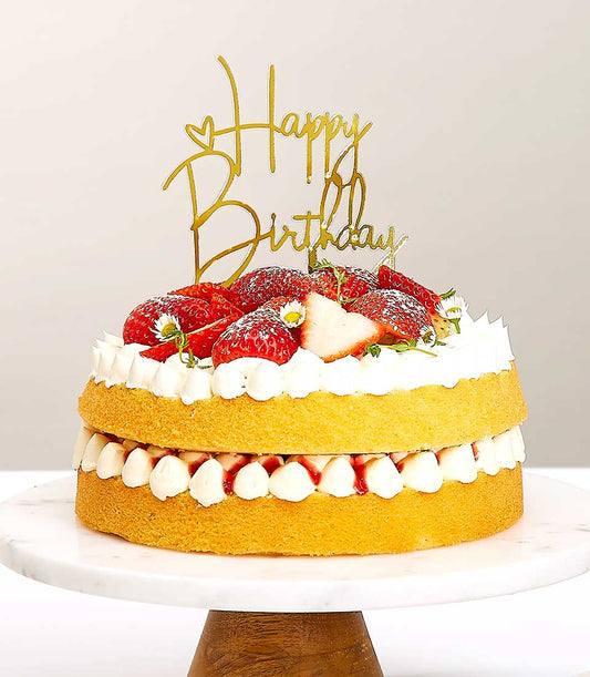 Two-tiered strawberry shortcake with fresh berries, whipped cream, and gold birthday topper on marble stand