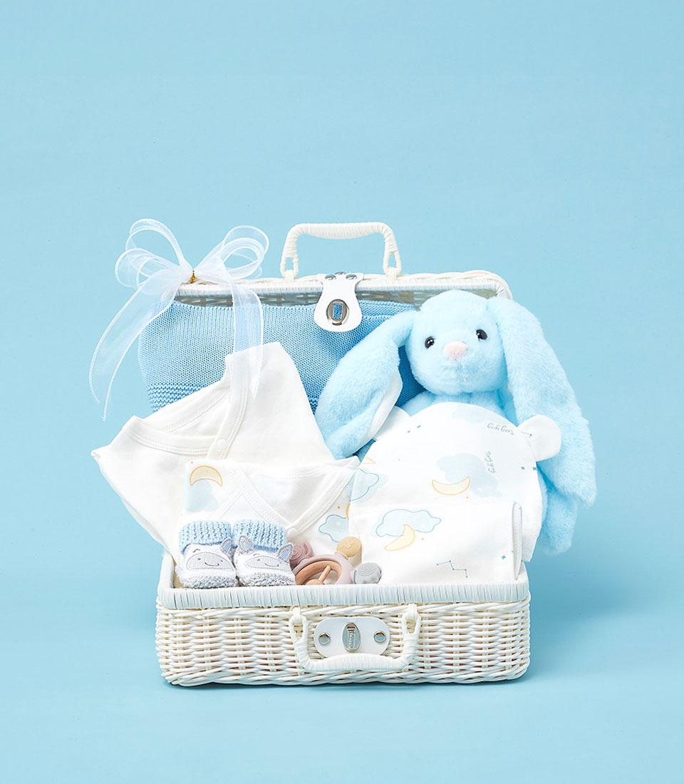 Elegant white wicker baby hamper filled with light blue blanket, onesie, moon-cloud swaddle, plush bunny, and booties for
