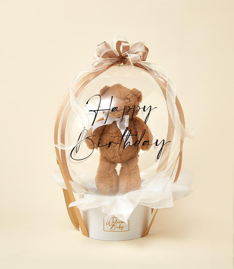 Brown teddy bear in clear birthday balloon with gold ribbon bow, white tissue base in cylindrical gift container