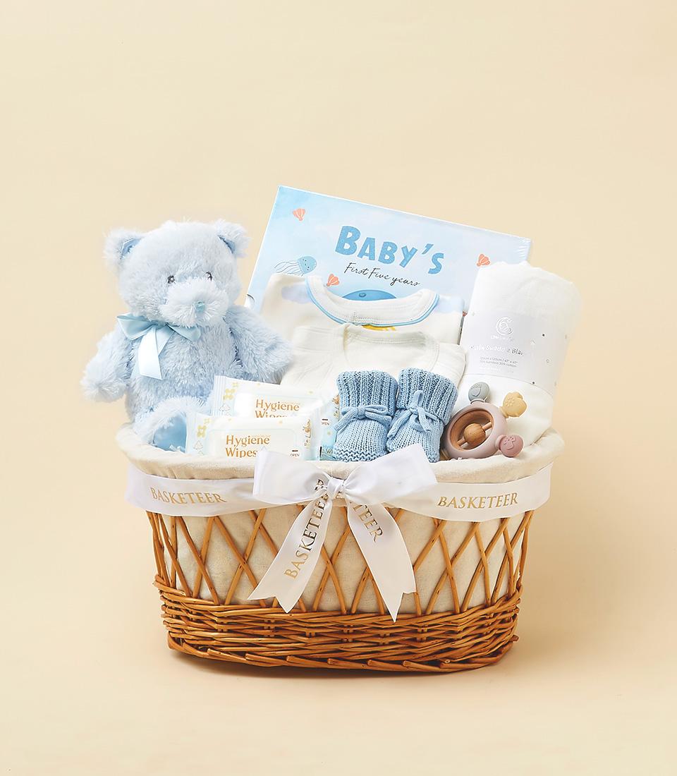 Blue and white newborn gift basket with teddy bear, memory book, bodysuits, and baby essentials in woven wicker