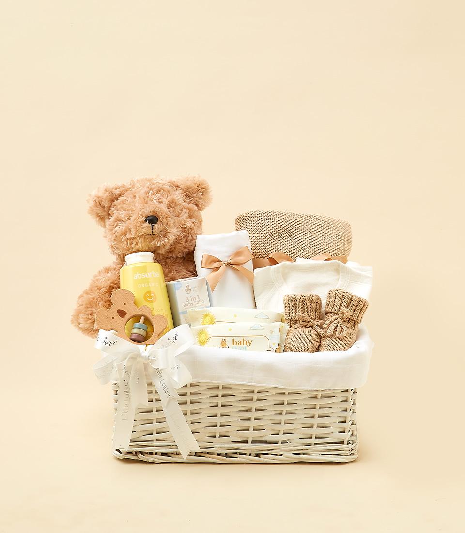 Newborn gift basket with plush teddy, organic baby bottle, wooden teether, balm, onesie, booties, and wipes in white wicker