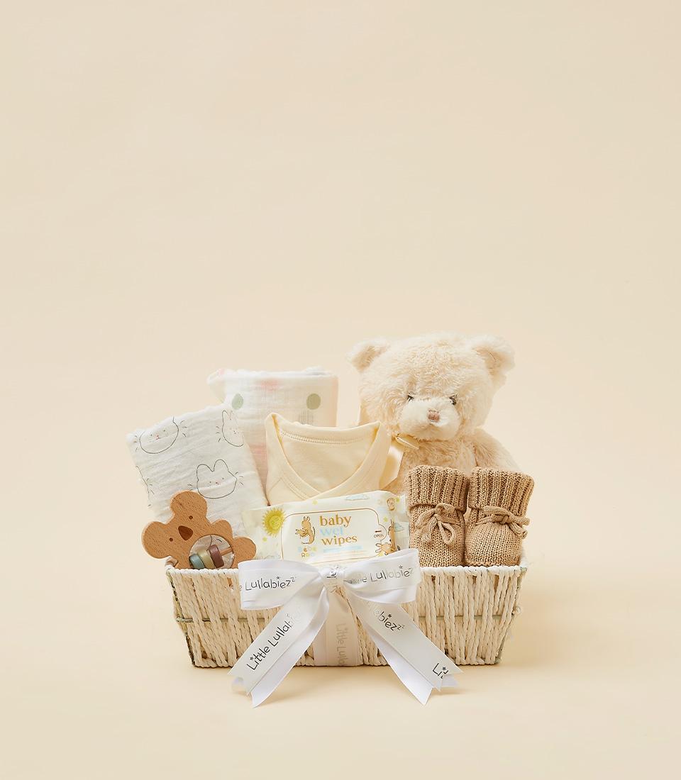 Woven baby gift basket with white muslin cloths, yellow onesie, wooden teether, plush teddy bear, and brown booties