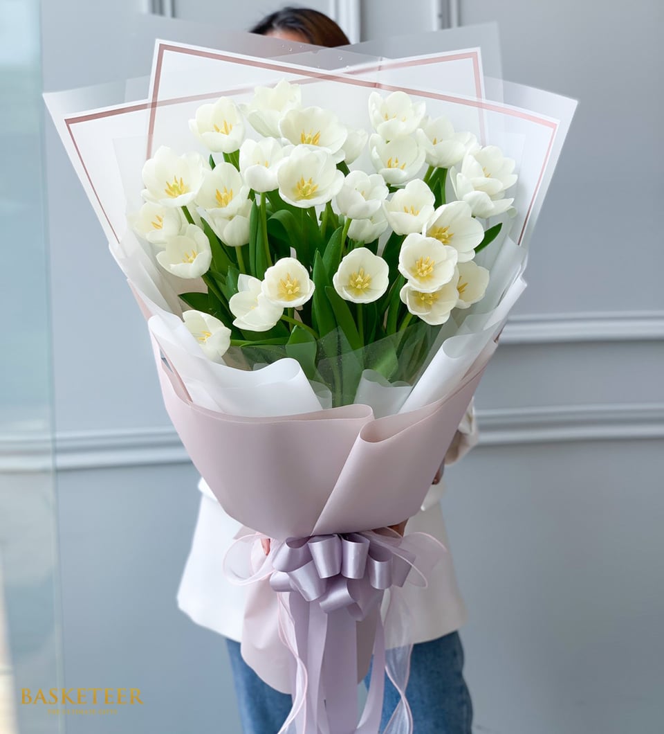 White tulips with yellow centers arranged in layered lavender and tissue wrapping, held against white top and blue jeans