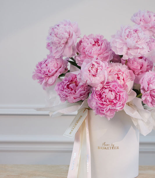 Pink peonies in a white box with cream ribbon, branded "Flowers by Basketeer" against a light gray wall.