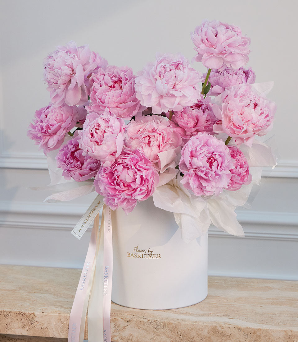 Pink peonies in a white ceramic vase with "Flowers by Basketeer" logo, tied with cream and pink ribbons, displayed against a white paneled background.