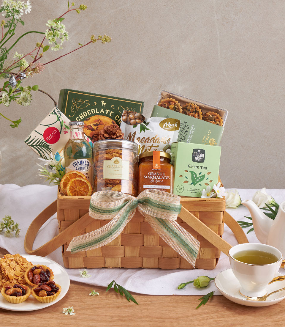 Woven gift basket with chocolate chip cookies, macadamia nuts, green tea, and gourmet spreads tied with green striped ribbon