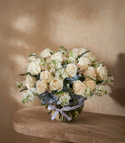 Cream roses and blue hydrangeas in clear glass vase with white ribbon bow