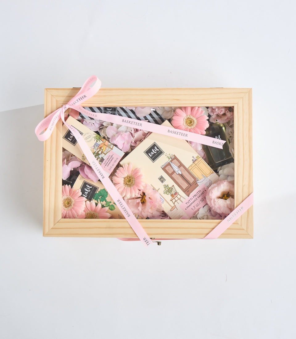 Luxury gift box with natural wood frame, pink ribbons, fresh pink and white flowers, cosmetic products, and zebra-print accents on white background.