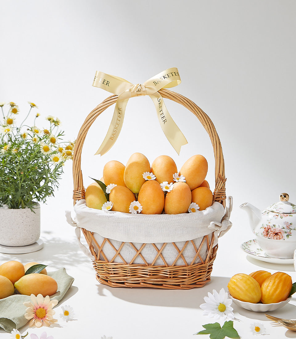 Woven wicker basket filled with ripe yellow mangoes and white daisies, tied with gold Basketeer ribbon