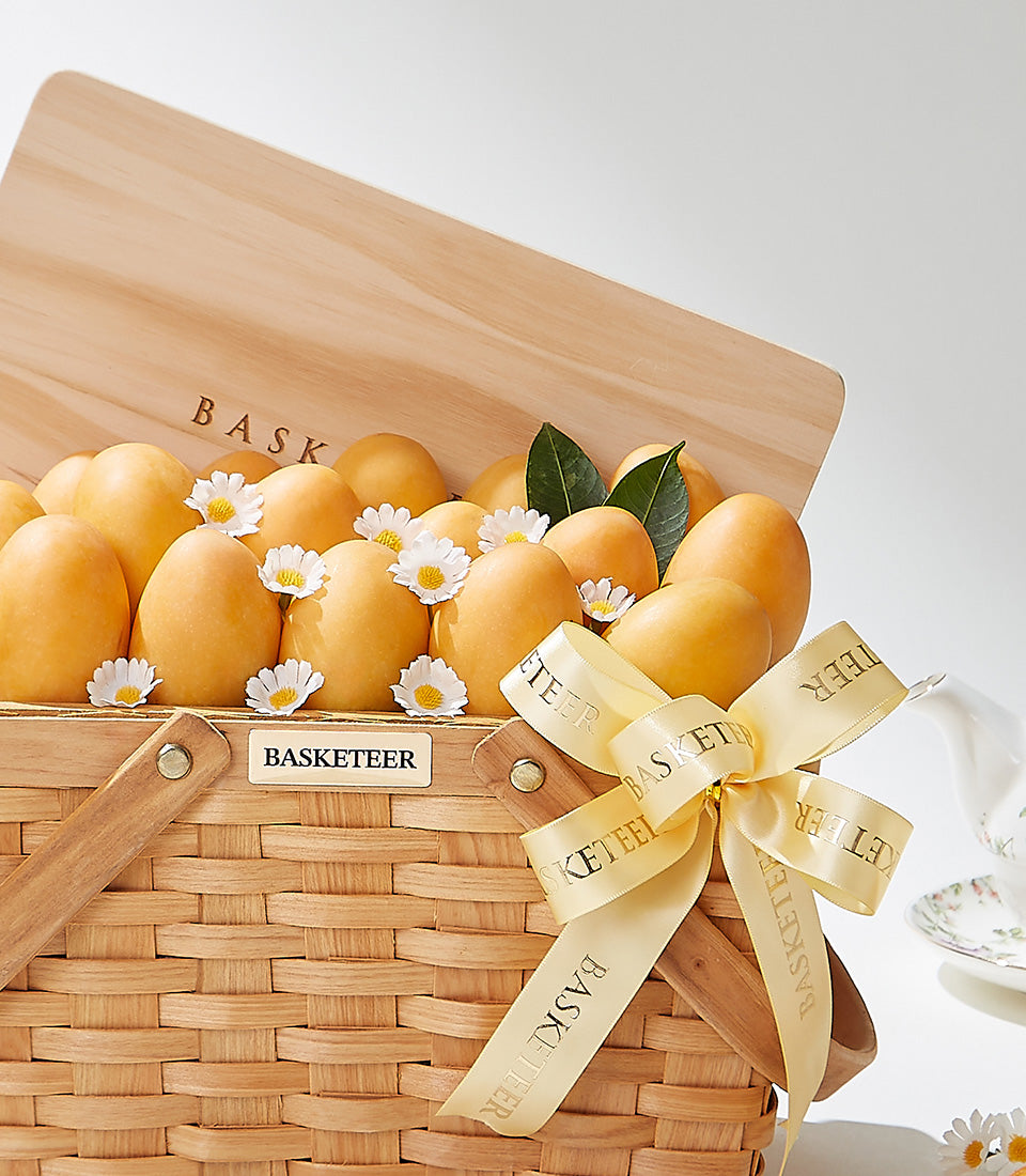 Premium woven basket filled with golden mangoes, white daisies, and fresh green leaves with gold ribbon bow