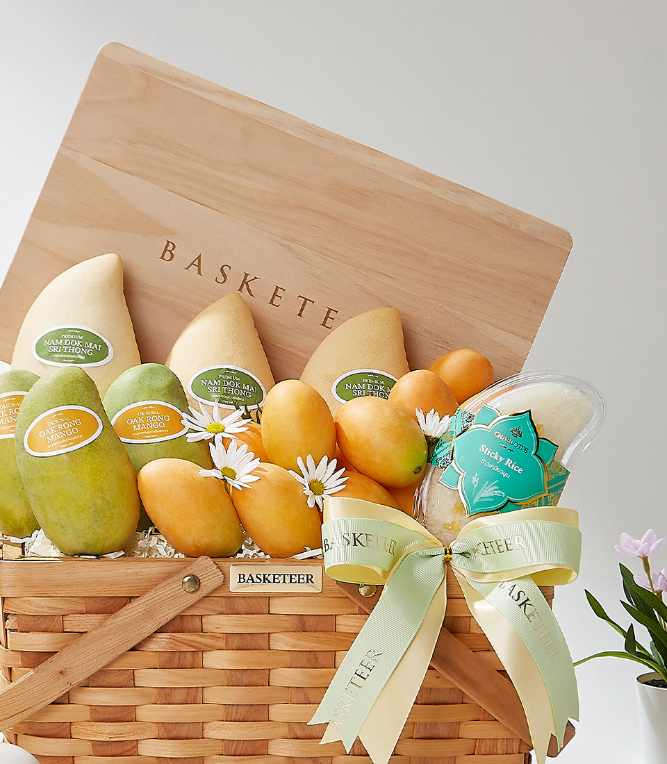 Golden woven gift basket with Thai mangoes, sticky rice, and white flowers, tied with cream and green ribbon