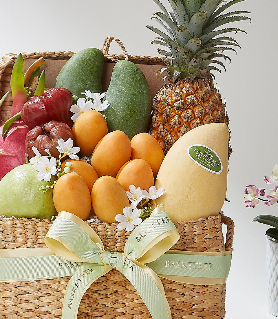 Tropical mixed fruit basket with mangoes, pineapple, dragon fruit, and rose apples in woven straw with green ribbon
