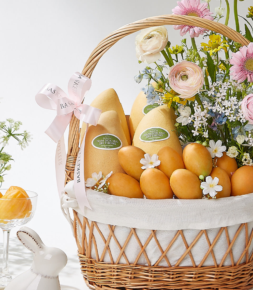 Woven wicker basket with Thai mangoes, tropical fruits, pink gerberas, and white ranunculus flowers with ribbon bow