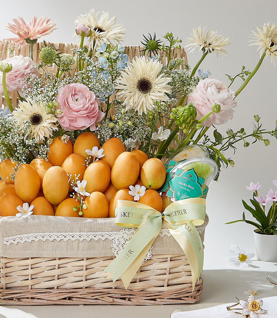 Deluxe Thai fruit and flower basket with golden mangoes, pink ranunculus, cream gerberas, and blue delphiniums