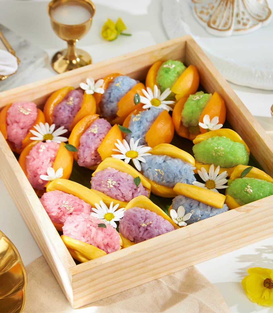 Colorful rainbow sticky rice desserts in tropical fruit shells with edible gold leaf and flowers on wooden tray