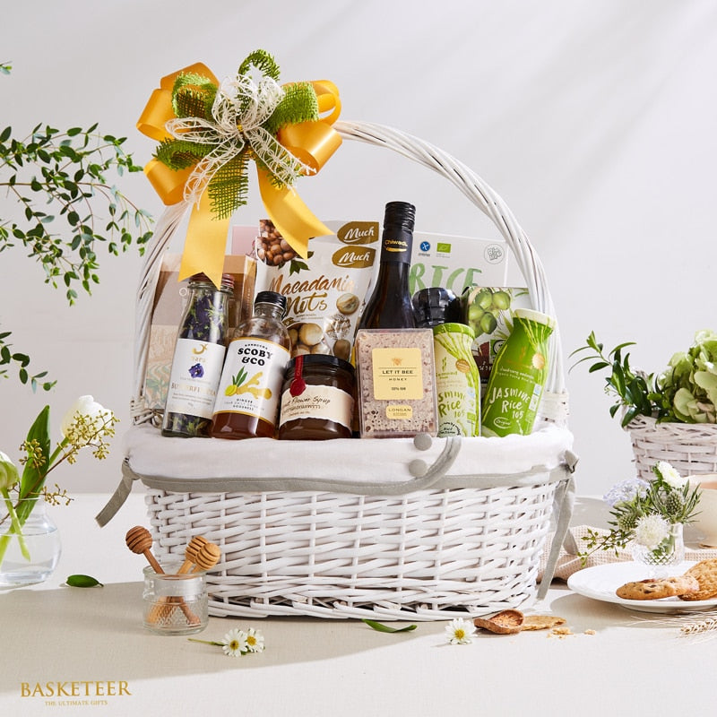 White wicker gift basket with gourmet Thai products, yellow-green bow, jasmine rice, honey, and artisan beverages
