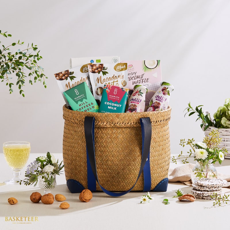 Woven straw gift basket with blue leather handles filled with macadamia, coconut waffle, hill coffee, and Thai gourmet treats