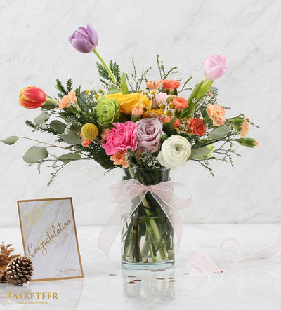 Vibrant mixed flower arrangement in clear glass vase with tulips, roses, and carnations, tied with pink ribbon
