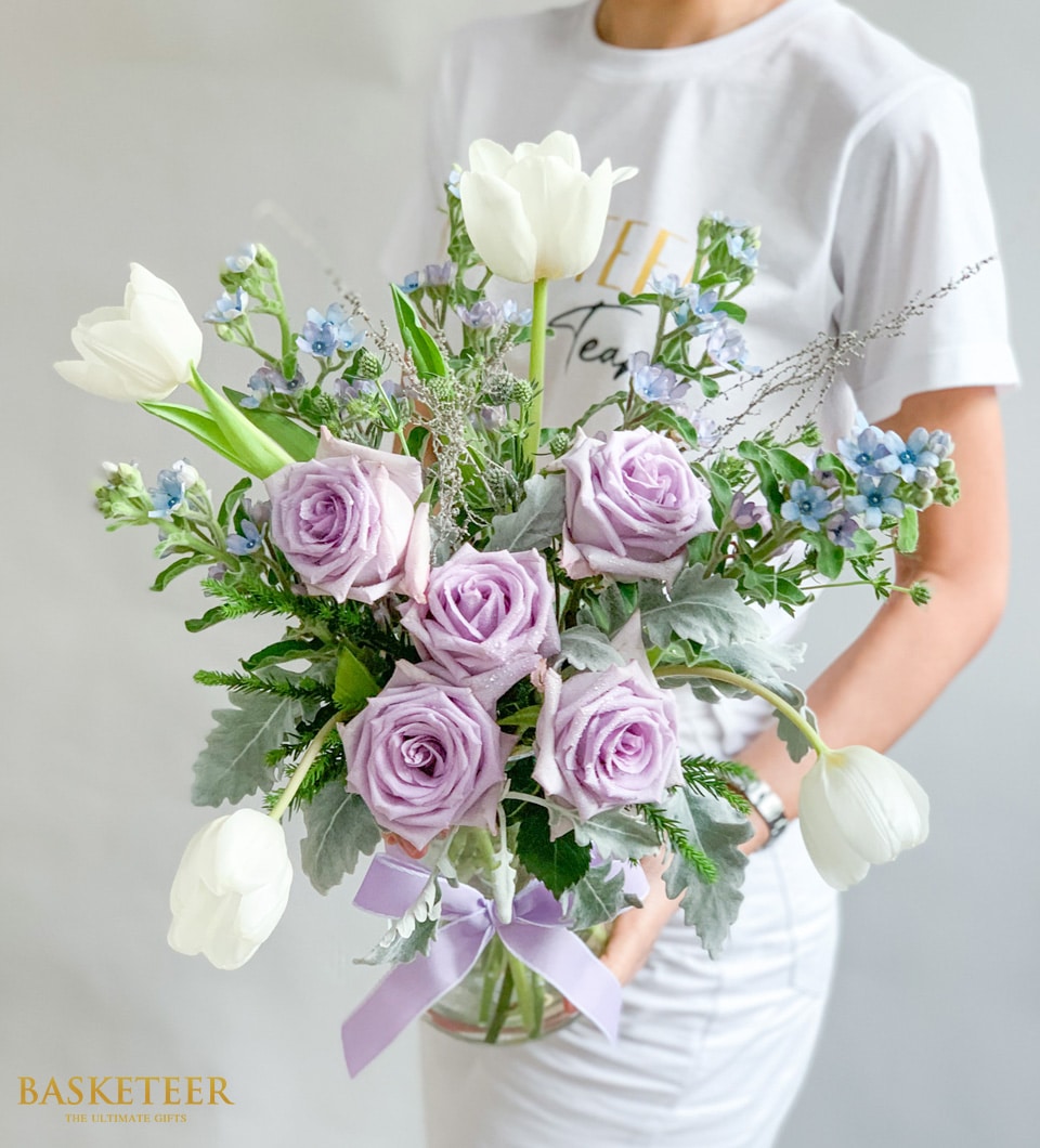 Purple roses and white tulips with dusty miller in clear glass vase, tied with lavender ribbon