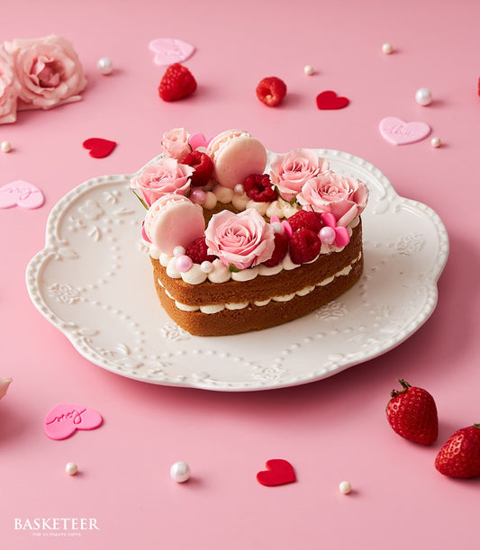 Heart-shaped vanilla cake with pink roses, macarons, and fresh raspberries on white scalloped plate