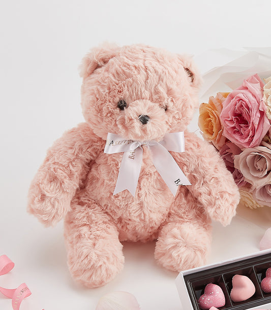 Pink teddy bear with white satin bow, fresh roses in blush and peach tones, and heart-shaped chocolates Valentine's gift