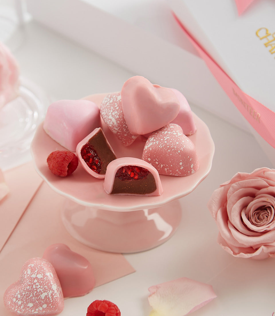 Pink raspberry passion heart-shaped bonbons with dark ganache filling, fresh raspberries, and rose petals on ceramic stand