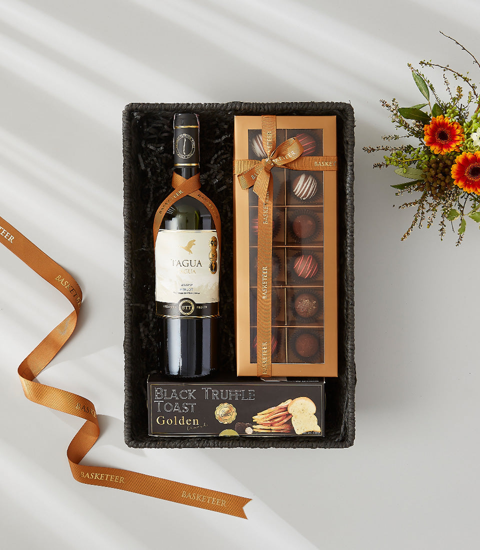 Premium wine gift basket with red wine, artisan chocolates, and gourmet crackers in black shredded paper, gold ribbon accents