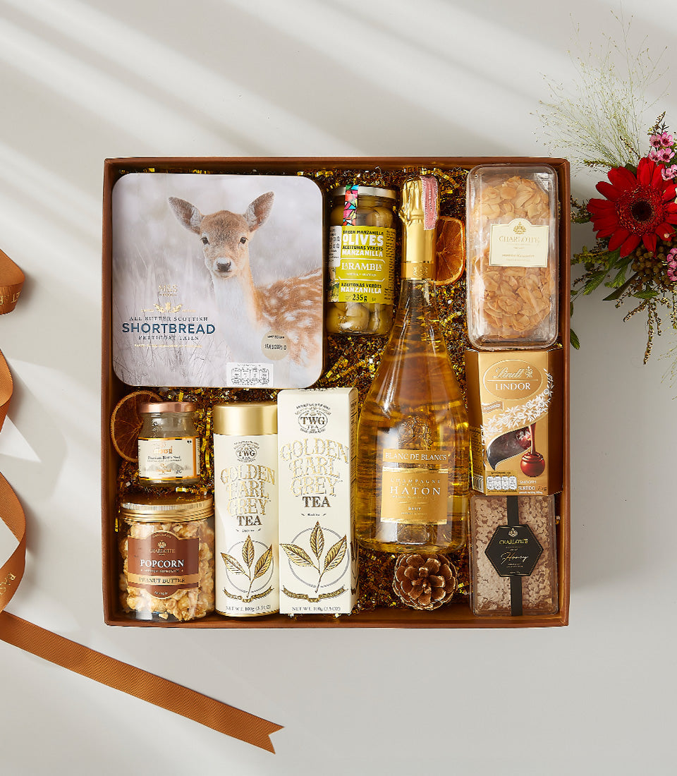 Luxury champagne gift hamper with champagne, shortbread, tea, olives, honey, and chocolate on golden shredded paper