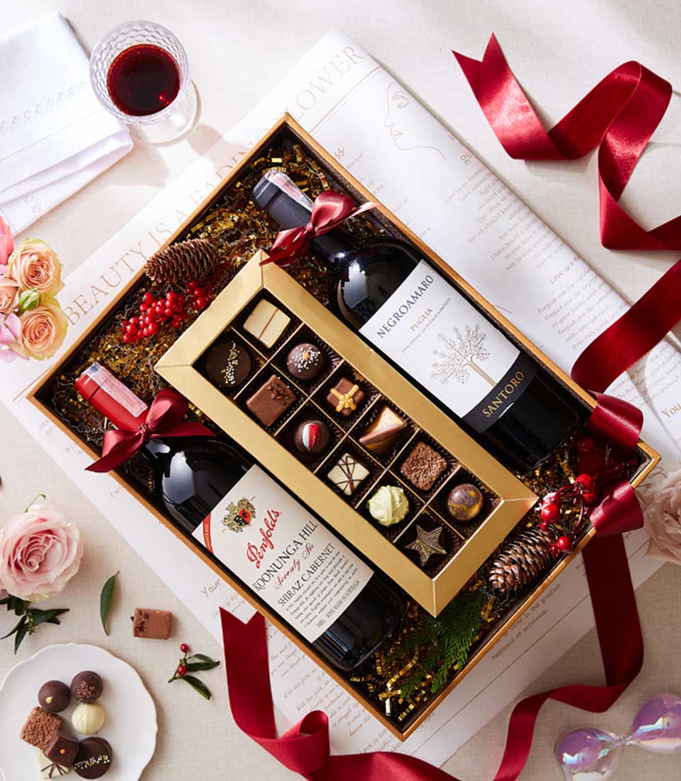 Luxury wine and chocolate gift box with two red wine bottles, artisan chocolates, and festive gold-shredded paper styling