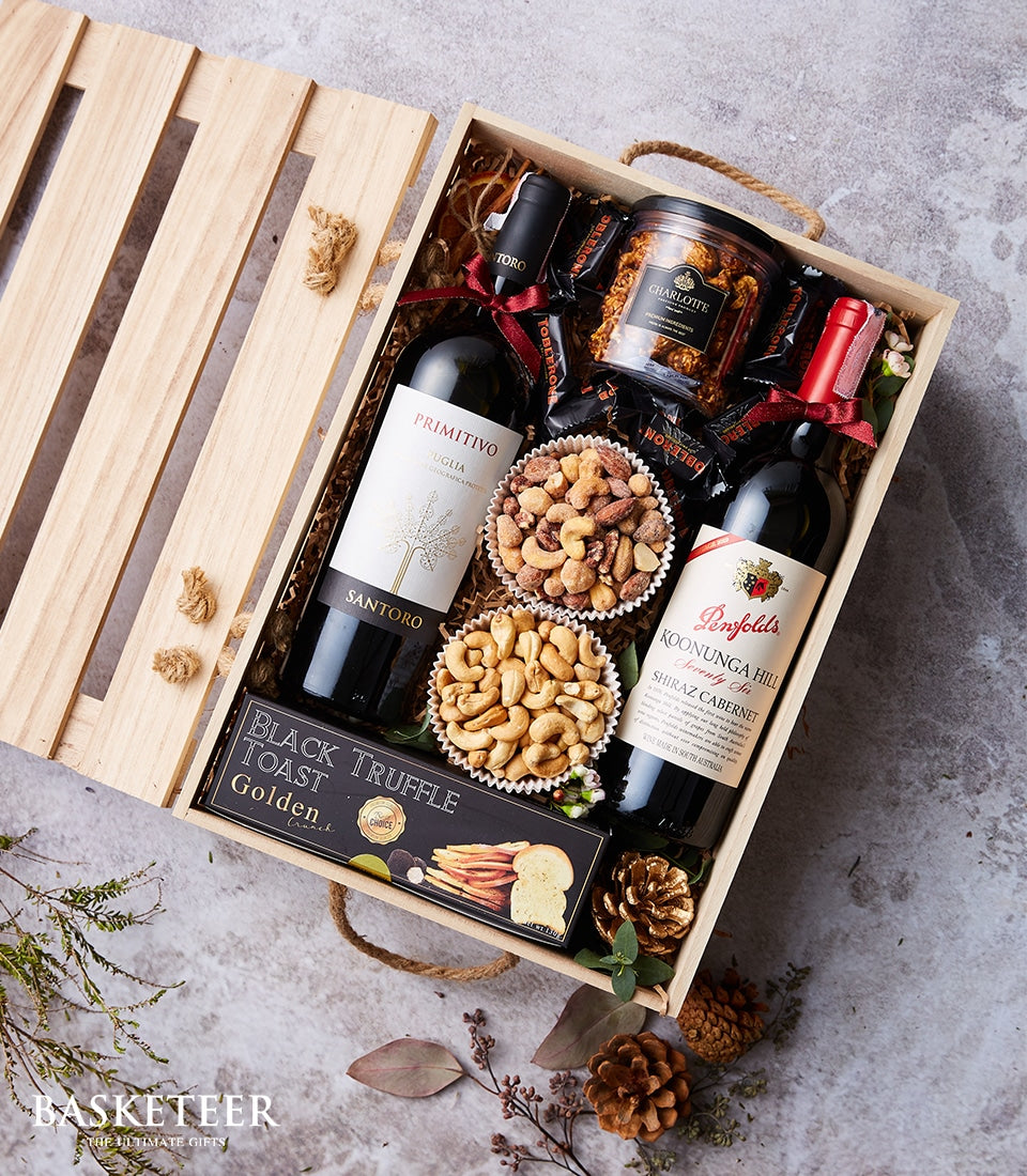 Wooden gift crate with red wine bottles, roasted nuts, dark chocolates, and gourmet snacks on shredded paper