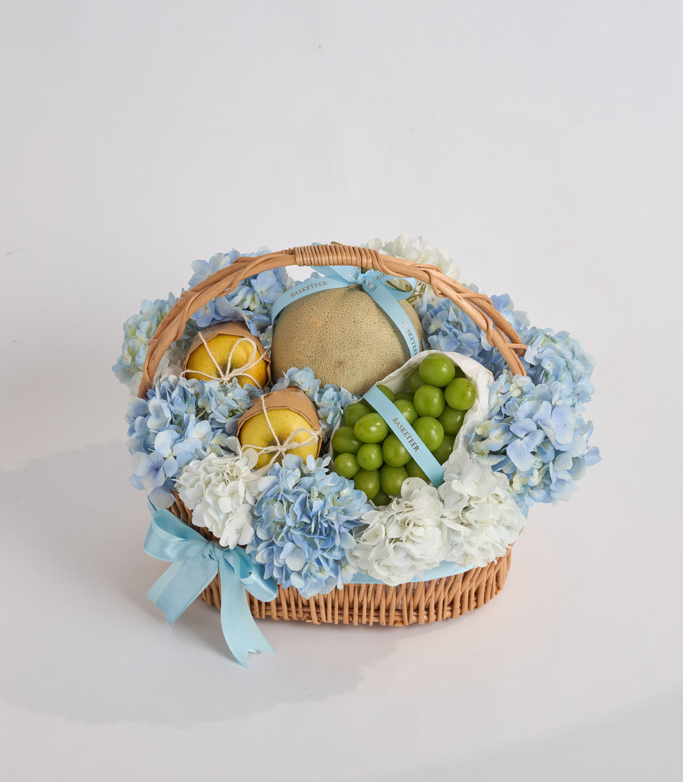 Wicker basket filled with blue and white hydrangeas, green grapes, sliced lemons, a melon, and light blue ribbons.